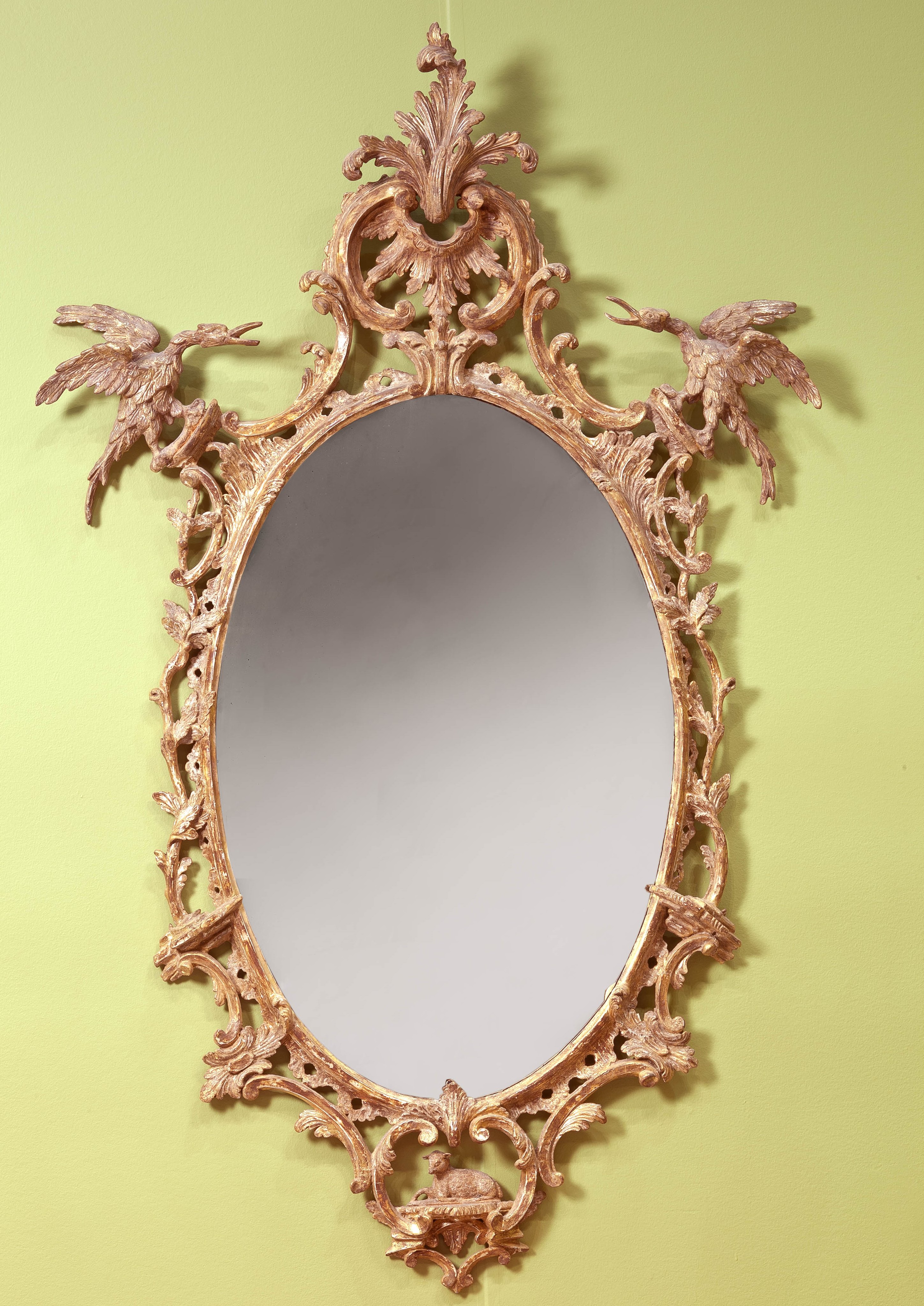 A fine and impressive oval rococo looking glass crested with a double c-scrolls holding an acanthus spray flanked by ho-ho birds over trailing vines with carved c-scrolls, the base terminating with c-scrolls flanking a seated lamb. Glass not original. English, Circa 1755. H. 52 ½,” W. 32 ¾.” Courtesy Clinton Howell Antiques.